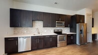 This well-designed single-floor residence offers a thoughtfully laid-out interior with contemporary finishes throughout. The kitchen is a standout feature, outfitted with rich dark-stained shaker-style cabinetry that extends to the ceiling, crisp white countertops, a textured stone tile backsplash, and a full suite of stainless steel appliances including a refrigerator with ice maker, electric range, over-the-range microwave, and dishwasher. Light hardwood-style flooring runs throughout the kitchen and main living areas, adding warmth to the space. The bedroom is bright and airy, featuring multiple windows that invite natural light, a ceiling fan with integrated lighting, and the same hardwood-style flooring for a cohesive look. The bathrooms are finished with neutral, large-format tile work and include quality fixtures. One bathroom offers a soaking tub with a tiled surround accented by a decorative mosaic border, while another features a separate walk-in shower with matching tile. A walk-in closet with built-in shelving provides ample storage. Additional details include a sliding barn-style door, a programmable thermostat, and clean white walls throughout, contributing to a modern and move-in-ready atmosphere.
