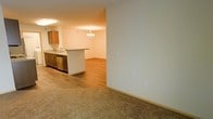 This single-floor residence offers a thoughtfully designed open-concept layout that seamlessly connects the kitchen and living areas. The kitchen is equipped with warm wood-toned cabinetry, a stainless steel dishwasher, and a light-toned countertop peninsula that doubles as a breakfast bar and natural divider between the cooking and living spaces. Hardwood-style flooring runs throughout the kitchen and dining area, providing a clean, modern aesthetic, while plush carpeting in the adjacent living area adds warmth and comfort underfoot. A decorative chandelier serves as a focal point, adding a touch of elegance to the dining or living space. The walls are finished in a neutral, warm tone that complements the overall color palette and creates a bright, welcoming ambiance. The open flow between spaces makes the layout feel expansive and versatile, well-suited for both everyday living and entertaining. Clean lines, practical finishes, and a cohesive design language give this single-level space a functional yet inviting character throughout.
