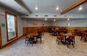 This single-floor gathering space is thoughtfully designed to accommodate a variety of events and group activities. The main hall is generously proportioned, featuring striking exposed wooden ceiling beams that add warmth and rustic character to the otherwise contemporary interior. Recessed lighting and polished concrete-style flooring run throughout, creating a clean, durable foundation. Multiple long wooden dining tables with cushioned chairs are arranged throughout, offering flexible seating configurations suited for large gatherings or smaller group settings. One section of the hall features warm knotty wood wainscoting along the lower walls, complemented by framed artwork, lending a cozy, lodge-like ambiance. A row of large wood-trimmed windows and glass doors along one wall floods the space with natural light and provides views of the surrounding outdoor area. A dedicated kitchen anchors one end of the space, fully equipped with stainless steel appliances including a professional-grade range with a large hood vent, a full-size refrigerator, dark cabinetry with subway tile backsplash, and a teal island with a quartz countertop. Industrial-style pendant lights illuminate the kitchen workspace. A corkboard and wall clock near a quieter dining alcove add functional, community-oriented touches throughout.