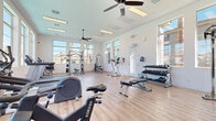 This single-floor fitness center is designed to support a comprehensive workout experience in a bright, welcoming environment. The space is defined by its clean white walls, light hardwood-style flooring, and an abundance of large windows that line multiple walls, flooding the room with natural light and offering views of the surrounding neighborhood, including palm trees and residential rooftops. Ceiling fans are strategically placed throughout to maintain comfortable airflow during workouts.

The layout is thoughtfully organized to accommodate both cardio and strength training. Cardio equipment includes treadmills—among them a Life Fitness model—a stationary bike (ESB250), an elliptical trainer, and a rowing machine. For strength and resistance training, the space offers a multi-station cable and pulley machine, a captain's chair for core work, a preacher curl bench, a flat utility bench, and a well-stocked dumbbell rack ranging across multiple weight increments. Resistance bands are also available for functional training.

Recessed and strip lighting supplements the natural light, ensuring visibility throughout the day and evening. Safety features include clearly marked exit signage and a fire extinguisher. The overall ambiance is clean, modern, and functional, making this fitness center a practical and inviting space for a wide range of fitness routines.