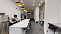 Located at 55 W Chestnut St in Chicago's Near North Side neighborhood, this single-floor amenity space is designed to serve as a dynamic communal hub with a refined, modern aesthetic. Soaring ceilings amplify the sense of openness throughout, while floor-to-ceiling windows along one wall invite generous natural light and offer views of the tree-lined street outside.

The kitchen area is fully equipped with stainless steel appliances—including a refrigerator, range, and microwave—complemented by sleek white cabinetry and a long white quartz island with bar-height seating. A striking arrangement of black-and-gold pendant lights cascades above the island, adding a bold decorative focal point.

Adjacent to the kitchen, a lounge zone features contemporary seating in teal and gray tones, arranged around structural columns that define the space without enclosing it. A large-scale decorative map of Chicago's waterways serves as a distinctive wall feature, grounding the space in its urban context. A shuffleboard table adds a recreational dimension nearby.

The entertainment area includes a wall-mounted flat-screen television with a media console, providing a comfortable gathering spot. Colorful abstract artwork adorns the walls, contributing vibrancy and personality. Throughout, gray carpet ties the zones together, creating a cohesive and inviting environment suited for socializing, dining, and recreation.