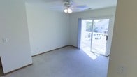 This single-floor space offers a clean, straightforward layout centered around a well-lit main living area. The room is finished with wall-to-wall carpeting in a neutral tone, providing a comfortable and cohesive feel underfoot. White walls and white baseboard trim keep the space feeling fresh and open.

A ceiling fan with an integrated light fixture is centrally mounted, offering both overhead illumination and air circulation year-round. A large sliding glass door serves as a prominent architectural feature, flooding the room with abundant natural light and providing direct access to an outdoor deck. The deck, framed with white railing, overlooks a residential neighborhood with mature trees visible in the background, adding a pleasant outdoor connection to the interior.

The overall ambiance is calm and understated, with a neutral color palette that lends versatility for a variety of furnishing styles. The simplicity of the space, combined with the natural light and outdoor access, creates an inviting and functional environment well-suited for everyday living.