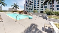 This rooftop amenity deck offers a thoughtfully designed outdoor retreat set against a dramatic urban skyline backdrop. At its centerpiece sits a generously sized lap pool with vivid turquoise water, flanked by a wide sundeck furnished with cushioned lounge chairs, woven pod-style seating cabanas, and market umbrellas—ideal for relaxing under open skies. Adjacent to the pool, a circular spa provides a dedicated space for hydrotherapy with city views stretching to the horizon. Tall palm trees line the perimeter, reinforcing a resort-inspired atmosphere. A separate outdoor lounge area features a large glass-enclosed fire pit table surrounded by plush sectional seating, accent stools, and rustic wood side tables, creating an inviting setting for evening gatherings. Raised planters throughout the deck are filled with drought-tolerant ornamental grasses, succulents, agaves, and flowering plants, contributing a lush, garden-like quality. A tiered grass lawn area with curved concrete steps offers an additional green space for casual use or lounging. Vibrant teal accent furniture pieces are scattered throughout, providing cohesive pops of color. The entire deck benefits from abundant natural light and panoramic views of the surrounding high-rise cityscape, making it a compelling urban outdoor amenity.