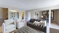 This well-designed two-floor space offers a cohesive blend of comfort and contemporary style. The living area serves as the heart of the home, showcasing a neutral color palette of soft grays and warm whites that creates a calm, inviting atmosphere. A dark upholstered sofa anchors the seating arrangement, complemented by plush cream accent chairs and a tufted gray ottoman that doubles as a coffee table. Built-in shelving along one wall provides both functional storage and an opportunity for decorative display, adding architectural character to the room. Elegant dual-lamp fixtures flank a sleek console table topped with tasteful greenery, while a large framed mirror reflects light and enhances the sense of openness. The carpeted flooring adds warmth underfoot, and the textured ceiling lends a classic finishing touch overhead. Doorways lead to additional rooms, including a bathroom visible from the main living space, hinting at a well-connected and functional floor plan. The overall ambiance is modern yet approachable, with carefully chosen furnishings and a refined neutral scheme that makes the space feel polished and livable across both levels.
