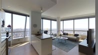 This sophisticated high-rise residence occupies a single floor and is defined by its open-concept layout, clean lines, and an abundance of natural light. Floor-to-ceiling windows wrap around the living and kitchen areas, framing breathtaking panoramic views of the cityscape that stretch to the horizon, making the skyline a constant visual centerpiece.

The kitchen is thoughtfully integrated into the main living space, featuring a central island with a dark countertop, an undermount sink, and sleek white cabinetry. Light hardwood flooring runs throughout, lending warmth to the otherwise contemporary aesthetic and tying the spaces together seamlessly.

The living area is positioned to take full advantage of the surrounding views, with generous natural light flooding the space throughout the day. The open-plan arrangement creates a sense of expansiveness, allowing the kitchen and living room to flow effortlessly into one another.

The overall ambiance is modern and refined, characterized by a neutral white palette, minimalist architectural details, and a bright, airy atmosphere. The combination of high elevation, wraparound windows, and clean contemporary design creates a living environment that feels both elevated and inviting.