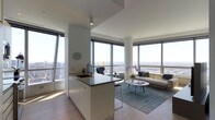 This sophisticated high-rise residence occupies a single floor and is defined by its open-concept layout, clean lines, and an abundance of natural light. Floor-to-ceiling windows wrap around the living and kitchen areas, framing breathtaking panoramic views of the cityscape that stretch to the horizon, making the skyline a constant visual centerpiece.

The kitchen is thoughtfully integrated into the main living space, featuring a central island with a dark countertop, an undermount sink, and sleek white cabinetry. Light hardwood flooring runs throughout, lending warmth to the otherwise contemporary aesthetic and tying the spaces together seamlessly.

The living area is positioned to take full advantage of the surrounding views, with generous natural light flooding the space throughout the day. The open-plan arrangement creates a sense of expansiveness, allowing the kitchen and living room to flow effortlessly into one another.

The overall ambiance is modern and refined, characterized by a neutral white palette, minimalist architectural details, and a bright, airy atmosphere. The combination of high elevation, wraparound windows, and clean contemporary design creates a living environment that feels both elevated and inviting.