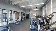 This fitness center offers a comprehensive workout environment designed to accommodate a wide range of exercise needs. The space is outfitted with an array of modern cardio equipment, including elliptical trainers and treadmills, neatly arranged along one side of the floor. On the opposite side, strength training machines provide options for targeted resistance workouts. The durable rubber flooring throughout the space is both practical and protective, ideal for high-traffic fitness use. The ceiling is open and industrial in style, featuring an exposed grid framework with integrated fluorescent lighting that ensures the space is brightly and evenly illuminated. Walls are finished in a cool grey tone, contributing to a clean and focused atmosphere. Large glass-paneled doors and interior windows allow for visibility between areas and bring in additional light, enhancing the sense of openness. A row of lockers is visible near the back of the space, providing convenient storage for users. The overall layout is spacious and well-organized, allowing for comfortable movement between equipment. This single-floor facility is thoughtfully designed to support a productive and motivating fitness experience.