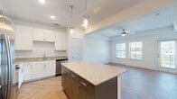 This single-floor residence showcases a thoughtfully designed open-concept layout where the kitchen and living area flow seamlessly together. The kitchen is a standout feature, outfitted with crisp white cabinetry extending to the ceiling, granite countertops, and a full suite of stainless steel appliances including a French door refrigerator, range, microwave, and dishwasher. A substantial central island with a dark base and granite surface provides additional prep space and casual seating. Pendant globe lights hang above the island, adding a modern touch. Throughout the main living areas, herringbone-patterned hardwood flooring creates a striking visual element underfoot. The generously sized living area is bright and open, featuring a ceiling fan, natural light from multiple windows, and direct access to an outdoor balcony. The bedroom is spacious and airy, also featuring herringbone flooring, a ceiling fan, and access to what appears to be a closet. The bathroom is finished with classic white subway tile surrounding the tub and shower, granite-topped white vanity cabinetry, and coordinating hardwood-style flooring, maintaining the cohesive, contemporary design found throughout the space.