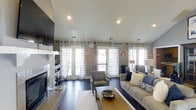 This single-story space showcases a well-appointed living area designed with both comfort and aesthetics in mind. The room is anchored by a tiled fireplace on one wall, accompanied by a wall-mounted flat-screen television, creating a functional focal point. Dark hardwood flooring runs throughout, providing a rich contrast to the light-toned walls and ceiling.

Natural light pours in abundantly through multiple large windows and a set of French doors, all dressed with coordinating navy and white patterned curtains that add a decorative touch while framing the outdoor views. Recessed lighting in the smooth white ceiling supplements the natural light, ensuring the space is well-lit throughout.

The seating arrangement includes a plush neutral sofa accented with navy and cream throw pillows, paired with coordinating accent chairs. A wooden coffee table with a metallic bowl centerpiece anchors the seating area, which is set upon a light-toned area rug. The overall ambiance is cohesive and inviting, blending transitional design elements with practical layout choices that make the space feel both polished and livable.