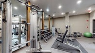 The fitness center at MAA Piedmont Park is a thoughtfully designed, single-level space that provides a comprehensive workout environment for residents. The room features a neutral color palette of light gray and white walls, with durable flooring and recessed lighting supplemented by ceiling fans to maintain a comfortable atmosphere during exercise.

The cardio area is equipped with treadmills, elliptical machines, and a stair climber, all positioned near frosted windows that allow natural light to filter in. Two wall-mounted flat-screen televisions are positioned for easy viewing from the cardio equipment. Rubber floor mats are placed throughout for safety and equipment protection.

The strength training zone is anchored by a cable machine with multiple attachment options and framed by full-length mirrors accented with warm wood trim columns, which visually expand the space. A well-organized dumbbell rack offers a range of free weights alongside adjustable benches and selectorized weight machines.

A dedicated stretching and functional training area houses stability balls, medicine balls on a vertical rack, and additional floor mats. The space also includes a wall clock and restroom access nearby, adding convenience. Overall, the fitness center offers a clean, modern, and well-rounded facility.