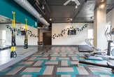 This fitness center in Atlanta, GA (30318) offers a comprehensive workout facility spread across a single floor, thoughtfully designed to accommodate a wide range of exercise needs. The space features an industrial-modern aesthetic, with exposed ductwork, concrete ceilings, and track lighting that lend an energetic, contemporary character throughout.

The main workout area is divided into distinct functional zones. The cardio section is lined with large windows that invite abundant natural light and provide views of the surrounding greenery, housing multiple treadmills, ellipticals, a stair climber, and a rowing machine. The strength training zone is well-appointed with free weights, a weight bench, cable machines, a squat rack, and a multi-station functional trainer, complemented by floor-to-ceiling mirrors that visually expand the space. A separate, more intimate room is dedicated to indoor cycling bikes, offering a focused environment for spin workouts.

Underfoot, a striking geometric patterned floor in neutral tones accented with teal ties the space together visually. Decorative wall art, including a flowing teal leaf sculpture and colorful canvas paintings, adds personality and warmth to the otherwise utilitarian setting. A wall-mounted television provides entertainment or informational content for users. Ceiling fans ensure comfortable airflow throughout, rounding out this thoughtfully designed and fully equipped fitness facility.