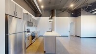 Located on University Drive North in Fargo, this two-bedroom, two-bathroom unit showcases a striking industrial-modern aesthetic throughout. Soaring ceilings with exposed ductwork, structural columns, and recessed lighting define the open-concept kitchen and living area, creating a loft-like atmosphere with impressive vertical scale. The kitchen is a standout feature, outfitted with light wood cabinetry, stainless steel appliances, a spacious center island with a built-in sink, and sleek pendant lighting overhead. Polished concrete floors flow seamlessly throughout the main living areas, reflecting natural light and reinforcing the unit's contemporary character. The generously sized primary bedroom offers ample room and benefits from large windows that bring in abundant natural light and street-level views. The second bedroom also features large windows and a structural column detail, adding architectural interest. Both bathrooms are finished with light wood vanities, white countertops, and polished concrete flooring. One bathroom includes a full tub and shower combination. Additional conveniences include dedicated closet storage in multiple areas and an in-unit laundry room, making this a well-appointed and functional urban living space.