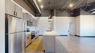 Located on University Drive North in Fargo, this two-bedroom, two-bathroom unit showcases a striking industrial-modern aesthetic throughout. Soaring ceilings with exposed ductwork, structural columns, and recessed lighting define the open-concept kitchen and living area, creating a loft-like atmosphere with impressive vertical scale. The kitchen is a standout feature, outfitted with light wood cabinetry, stainless steel appliances, a spacious center island with a built-in sink, and sleek pendant lighting overhead. Polished concrete floors flow seamlessly throughout the main living areas, reflecting natural light and reinforcing the unit's contemporary character. The generously sized primary bedroom offers ample room and benefits from large windows that bring in abundant natural light and street-level views. The second bedroom also features large windows and a structural column detail, adding architectural interest. Both bathrooms are finished with light wood vanities, white countertops, and polished concrete flooring. One bathroom includes a full tub and shower combination. Additional conveniences include dedicated closet storage in multiple areas and an in-unit laundry room, making this a well-appointed and functional urban living space.