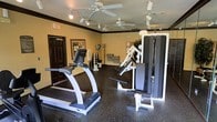 This fitness room offers a practical and functional space designed to support a variety of workout routines. The room is finished with durable rubber flooring in a dark speckled pattern, providing both safety and comfort during exercise. The walls are painted in a warm neutral tone, accented by dark crown molding that adds a clean, defined border between the walls and ceiling.

Overhead, multiple ceiling fans with integrated lighting ensure effective air circulation and a comfortable environment during workouts. Track lighting supplements the ceiling fans, providing well-distributed illumination throughout the space.

The room is equipped with a treadmill for cardiovascular training, positioned for easy access near the entrance. A multi-function cable weight machine anchors the strength training area, offering versatile resistance exercise options. A rack of dumbbells lines one wall, catering to free weight training needs. Additional equipment and storage elements are visible along the perimeter, maximizing the usable floor space in the center of the room.

A dark-framed door provides access to the space, and an exit sign is clearly posted for safety compliance. Overall, the fitness room is thoughtfully arranged to provide a comprehensive exercise environment.