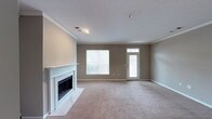This two-floor home offers a welcoming layout anchored by a spacious main-level living room. The room is finished with soft wall-to-wall carpeting in a neutral tone, complemented by warm greige walls that create a calm, understated atmosphere. A white-mantled fireplace with a traditional surround serves as the focal point of the room, adding a classic architectural detail to the space. Crown molding runs along the ceiling perimeter, lending a refined, finished look throughout. Natural light enters through a large front-facing window and a paneled entry door with a transom light above, brightening the interior without overwhelming it. The textured ceiling and recessed light fixture add subtle depth overhead. The open floor plan of the living area provides flexibility in furniture arrangement and transitions naturally to other areas of the home. With its combination of traditional design elements—crown molding, a fireplace surround, and neutral finishes—the main level establishes a cohesive and comfortable aesthetic. The home extends across two floors, offering additional living and private spaces beyond what is visible in the main living area.