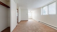 This single-floor residential unit offers a straightforward and functional layout designed for comfortable everyday living. The living area is generously proportioned, featuring warm beige wall-to-wall carpeting, crisp white walls, and a textured ceiling. Multiple windows dressed with horizontal blinds allow natural light to filter in, creating a bright and airy atmosphere. A ceiling fan provides additional air circulation, and baseboard heating runs along the perimeter walls throughout the unit, ensuring efficient warmth. A wall-mounted air conditioning unit in the living area offers year-round climate control.

The bedrooms are well-sized and carpeted, each featuring sliding closet doors that provide practical storage solutions. Warm wood-toned trim and door frames add a subtle contrast to the neutral color palette throughout.

The kitchen is compact and efficiently arranged, featuring warm wood cabinetry, a stainless steel sink, tile flooring, and included appliances such as a full-size refrigerator and electric range, making it move-in ready.

The bathroom is clean and classic in design, showcasing white square wall tiles surrounding a full bathtub and shower combination, complemented by mosaic tile flooring. The overall ambiance of the unit is light, neutral, and ready for personalization.