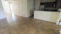 This single-floor space offers a clean, functional layout centered around an open-concept design that connects the living area to the kitchen. The floors throughout are finished with large-format neutral-toned ceramic or porcelain tiles, lending a cohesive and easy-to-maintain aesthetic across the space. The kitchen features dark cabinetry, a light-colored countertop with a peninsula-style overhang, and a stainless steel refrigerator, creating a practical and modern cooking area. A floor vent integrated into the baseboard area indicates a central HVAC system for climate control. The walls are painted in a soft, neutral tone, contributing to a bright and airy atmosphere. Multiple interior doors are visible, suggesting access to additional rooms or storage areas, including what appears to be a set of double doors that may serve as a closet or utility space. The overall layout is straightforward and well-organized, with clean lines and minimal ornamentation, making it a versatile space adaptable to a variety of uses. The open floor plan allows for natural sightlines between the living and kitchen areas, enhancing the sense of openness throughout.