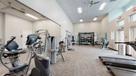 Located on 112th Ave N in Saint Petersburg, FL, this fitness center offers a comprehensive, single-floor facility designed to support a wide range of workout routines. The space features soaring ceilings throughout, creating an open and airy atmosphere that enhances the overall experience.

The main workout floor is generously sized, housing an extensive collection of strength training equipment, including multi-functional cable machines, a power rack, free weights, benches, and a variety of selectorized machines from brands such as Octane and Hoist. Full-length wall mirrors provide visual feedback and contribute to a sense of spaciousness.

A separate cardio area near the front entrance is bright and welcoming, with large windows and clerestory glazing flooding the space with natural light and views of the outdoors. Treadmills, ellipticals, and other cardio equipment are arranged here alongside wall-mounted TVs. A bold teal and neutral color-blocked accent wall adds a modern, energetic visual element.

An enclosed studio room with glass doors offers a more intimate setting, equipped with additional training equipment and mirrored walls, ideal for focused sessions or small group training. A dedicated hydration station with a water fountain is conveniently positioned nearby. Ceiling fans and panel lighting ensure comfort throughout. The facility is finished with durable carpet tile flooring suited for a high-traffic fitness environment.