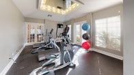 This fitness center is thoughtfully laid out across two interconnected rooms, offering a comprehensive workout environment for a wide range of training needs. The first room is dedicated to cardio and functional training, featuring multiple Star Trac elliptical machines and treadmills arranged beneath a striking tray ceiling with decorative lighting. French doors and large windows with white blinds flood the space with natural light, creating an open and energizing atmosphere. A wall-mounted television provides entertainment during workouts, and a stability ball rack adds a functional training element.

The second room transitions seamlessly into a strength and free weights area. A full-length wall mirror expands the sense of space and allows for form checks during exercise. This section houses a cable and pulley machine, a complete dumbbell rack, a weight bench, medicine ball storage, and additional cable resistance equipment. Ceiling fans with light fixtures provide comfortable airflow throughout both rooms. The durable rubber flooring throughout the facility offers both safety and sound absorption. Together, the two rooms form a cohesive, well-organized fitness facility that balances cardio, strength, and functional training in a bright, inviting setting.