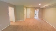 This single-floor space offers an open and functional layout highlighted by soft neutral wall tones in sage green and warm beige. The main living area is finished with plush wall-to-wall carpeting in a light tan hue, contributing to a comfortable and understated atmosphere. Recessed ceiling lights provide consistent overhead illumination throughout the space.

The open floor plan allows for a clear sightline from the main living area toward the kitchen, which is accessible through a wide doorway. The kitchen features light wood cabinetry, white appliances including a refrigerator and electric range, and tile or vinyl flooring that contrasts with the carpeted living space. The cabinetry provides ample storage, and the layout appears designed for practical everyday use.

Two interior doors are visible within the main living area — one leading to what appears to be a closet or utility space, and another providing access to an adjacent room or hallway. White trim and baseboards frame each doorway and run along the perimeter of the walls, adding a clean finishing detail throughout. Overall, the space presents a straightforward, move-in-ready environment with a cohesive and neutral aesthetic.