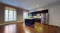This well-appointed single-floor urban residence offers a thoughtfully designed layout that balances style and functionality. The open-concept living and kitchen area serves as the heart of the space, anchored by rich, dark hardwood flooring that extends throughout. The kitchen is a standout feature, showcasing espresso-toned cabinetry, striking granite countertops, and a bold striped tile backsplash. A generously sized center island with pendant lighting overhead provides additional prep space and a natural gathering point. Stainless steel appliances, including a French door refrigerator, built-in microwave, and range, complete the chef-ready setup. Crown molding along the ceiling adds an elegant architectural touch to the living area, complementing the clean white entryway door and neutral wall tones. The bedroom is finished with plush wall-to-wall carpet and features a large window framing urban cityscape views, filling the room with natural light. The bathroom continues the refined aesthetic with tile flooring, granite-topped vanity cabinetry, and a bright window overlooking the city skyline. Throughout the residence, large windows invite abundant natural light and offer dynamic views of the surrounding urban environment, creating a vibrant and connected living experience.