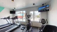Located at 195 E Main St in the heart of Columbus, Ohio, this modern fitness center offers a comprehensive and well-appointed workout environment across a single floor. The space is thoughtfully organized to accommodate a wide range of exercise disciplines, from cardio training to strength work and functional fitness.

Large windows line the cardio area, flooding the space with natural light and providing inspiring views of the Columbus downtown skyline. Multiple Spirit treadmills and an elliptical trainer are positioned to take full advantage of these city vistas, while wall-mounted televisions add an additional entertainment option during workouts.

The strength training section is equally well-equipped, featuring a multi-station cable machine, recumbent and upright bikes, and a dedicated free weights area with a full rack of dumbbells and kettlebells. A speed bag, adjustable weight bench, medicine balls, resistance bands, and battle ropes round out the functional training offerings, catering to a variety of fitness routines.

A bold teal and white geometric accent wall energizes the space visually, complementing the clean white walls and durable rubberized flooring throughout. Recessed and track lighting provide consistent illumination across all workout zones. The overall ambiance is clean, modern, and motivating — a fully equipped urban fitness facility designed to support diverse training needs.