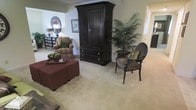 This single-floor space showcases a well-appointed living area characterized by neutral, wall-to-wall carpeting that lends a warm and comfortable feel throughout. The room is anchored by a large, dark-stained wood armoire, which serves as a prominent focal point and provides substantial storage. Seating options include a plaid upholstered armchair with green accent pillows and a distinctively patterned leopard-print accent chair with dark wooden legs, adding visual interest and texture to the space. A deep burgundy ottoman set serves as a functional coffee table centerpiece, styled with a decorative tray holding a bowl of greenery. A tall, lush indoor palm plant introduces a natural, organic element alongside the dark furnishings. Wall décor includes a round ornate mirror and a framed botanical print, contributing to the classical aesthetic. A dark mirrored console table near the entrance adds storage while complementing the room's color palette. Soft, neutral wall tones create a calm backdrop that allows the darker furniture pieces to stand out. A hallway visible in the background suggests additional rooms, hinting at a practical and well-organized floor plan throughout the residence.