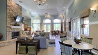 This inviting single-floor clubhouse space is thoughtfully designed to provide both comfort and elegance for its residents. The open-concept layout seamlessly blends multiple functional areas, creating a cohesive and welcoming environment throughout.

The main lounge area is furnished with plush gray sofas, upholstered ottomans, and accent seating arranged to encourage conversation and relaxation. Soft sage-green walls, accented by crisp white crown molding and chair rail detailing, lend a calm and refined atmosphere to the interior. Overhead, decorative chandeliers with wrought-iron frames serve as stylish focal points, complementing the recessed lighting throughout.

Soaring ceilings amplify the sense of openness, while a row of tall arched windows and glass-paned French doors along the rear wall bathe the space in abundant natural light and offer pleasant views of the surrounding greenery outside. Light-toned ceramic tile flooring runs consistently throughout, adding to the airy, clean aesthetic.

A dedicated dining area sits adjacent to the lounge, furnished with round and rectangular tables and mixed seating, ideal for casual gatherings. A kitchenette or bar area is visible toward one end of the space, complete with counter seating and cabinetry, offering additional convenience. The overall ambiance is warm, polished, and well-appointed.