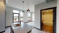 Located at 500 Bridge St NW in Grand Rapids, Michigan, this single-floor unit showcases a contemporary urban design with an open-concept layout and high-end finishes throughout. The kitchen is a standout feature, boasting dark cabinetry, stainless steel appliances, a white subway tile backsplash, and a central island with a quartz countertop and undermount sink — all finished with warm Edison bulb pendant lighting that adds an industrial character. Exposed ductwork along the ceiling enhances the loft-like aesthetic. Light wood-style flooring flows consistently throughout the space, complementing the clean, neutral color palette. The living area is generously proportioned, bathed in natural light from large floor-to-ceiling and double-pane windows that frame views of the surrounding Grand Rapids cityscape. The bathroom is well-appointed with dark wood cabinetry, a quartz vanity, an undermount sink, a full tub-shower combination, and industrial-style wall sconce lighting. Dark-framed interior doors and black hardware accents provide a cohesive, modern aesthetic that ties the space together. This unit offers a refined blend of industrial and contemporary design elements in a vibrant urban setting.