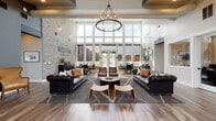 Located at 3807 SW Orbit St in Beaverton, Oregon, the West End District leasing and amenity space delivers a striking first impression through its dramatic double-height ceilings and expansive floor-to-ceiling windows that flood the interior with natural light and offer views of the outdoor pool area beyond. The single-floor layout is anchored by a spacious central lounge featuring rich dark leather Chesterfield sofas, cognac leather armchairs, and a bold round chandelier suspended from the soaring ceiling above. Wide-plank hardwood floors in a herringbone pattern add warmth and texture throughout. An exposed light brick accent wall proudly displays the "West End District" branding, lending an industrial-chic character to the space. A dedicated "Luxe Plans" wall showcases framed floor plan layouts alongside a console and dining table, serving as an informative consultation area for prospective residents. A private office or meeting room with glass partitions sits adjacent to the main lounge for one-on-one discussions. The palette of slate gray, crisp white shiplap walls, and natural wood tones creates a cohesive, contemporary aesthetic. A branded cruiser bicycle adds a playful, community-focused touch to the overall design.