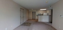 Located at 6785 Oakton Ln in Columbus, Ohio, this single-floor residence offers a clean and functional layout well-suited for everyday living. The main living area is carpeted and open, providing ample room to arrange furnishings freely. Natural light enters through the front entrance, which features a classic six-panel door with a tiled entryway mat area, creating a defined transition from the outdoors. The space flows seamlessly into a compact kitchen, which is separated from the living area by a raised countertop bar that doubles as a casual dining or workspace surface. The kitchen is equipped with white cabinetry offering generous storage, a range with overhead ventilation, a refrigerator, and under-counter appliances — all set against neutral-toned walls and flooring. A closet with sliding doors is positioned adjacent to the kitchen area, providing convenient built-in storage. The overall color palette throughout the space is neutral, with beige walls and light carpeting that create a calm, understated ambiance. The layout is straightforward and practical, making efficient use of the available space while maintaining an open and airy feel throughout.