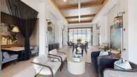 Located at 1124 Shoreview Cir in Casselberry, Florida, this single-story clubhouse is a showcase of contemporary design and resort-style living. The space greets visitors through dramatic floor-to-ceiling glass entry doors that flood the interior with abundant natural light and frame views of the surrounding grounds. Inside, soaring ceilings with richly detailed coffered wood inlays lend warmth and architectural depth to the open-plan layout, while large-format tile flooring flows seamlessly throughout. The main lounge area features a curated arrangement of stylish seating, including plush sofas and wooden accent chairs, centered around a round stone coffee table — creating an inviting space for gathering and conversation. A communal high-top table with bar stools serves as a versatile work and social hub, anchored by bold abstract artwork and lush greenery. Expansive steel-framed glass walls open the rear of the space to breathtaking views of a resort-style pool and outdoor terrace. A kitchenette area with custom cabinetry, open shelving, and patterned tile flooring offers convenience for events and everyday use. A dedicated office or leasing area provides a more private setting within the otherwise open layout. The overall ambiance is sophisticated yet welcoming, blending natural materials with modern finishes throughout.