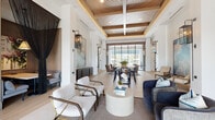 Located at 1124 Shoreview Cir in Casselberry, Florida, this single-story clubhouse is a showcase of contemporary design and resort-style living. The space greets visitors through dramatic floor-to-ceiling glass entry doors that flood the interior with abundant natural light and frame views of the surrounding grounds. Inside, soaring ceilings with richly detailed coffered wood inlays lend warmth and architectural depth to the open-plan layout, while large-format tile flooring flows seamlessly throughout. The main lounge area features a curated arrangement of stylish seating, including plush sofas and wooden accent chairs, centered around a round stone coffee table — creating an inviting space for gathering and conversation. A communal high-top table with bar stools serves as a versatile work and social hub, anchored by bold abstract artwork and lush greenery. Expansive steel-framed glass walls open the rear of the space to breathtaking views of a resort-style pool and outdoor terrace. A kitchenette area with custom cabinetry, open shelving, and patterned tile flooring offers convenience for events and everyday use. A dedicated office or leasing area provides a more private setting within the otherwise open layout. The overall ambiance is sophisticated yet welcoming, blending natural materials with modern finishes throughout.