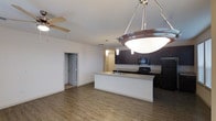 This single-floor space in Fort Worth showcases an open-concept design that seamlessly connects the living and kitchen areas. The kitchen is a standout feature, outfitted with dark espresso cabinetry, black appliances including a range and refrigerator, and a tile backsplash that adds texture to the space. A center island with a light countertop and an integrated sink provides both prep space and a natural dividing point between the kitchen and living area. Overhead, a large bowl pendant light anchors the kitchen zone, while additional pendant lights illuminate the island. The living area benefits from a ceiling fan and smooth white walls that keep the atmosphere bright and airy. Wood-look plank flooring runs consistently throughout, adding warmth and visual continuity. A doorway visible from the main living space leads to additional rooms, suggesting a functional and well-organized floor plan. The overall aesthetic is clean and contemporary, with a neutral palette complemented by the rich tones of the dark cabinetry, making the space versatile and move-in ready.