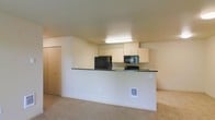 This single-floor space features an open-concept layout that integrates the kitchen and living area into one cohesive environment. The kitchen is positioned behind a half-wall peninsula with a dark countertop that serves as both a workspace and a visual divider between the two areas. Light maple cabinetry lines the upper wall, providing ample storage, and is complemented by black appliances, including a refrigerator, built-in microwave, and stovetop. A fluorescent ceiling fixture provides direct overhead lighting above the kitchen workspace. The walls throughout are finished in a warm neutral tone, contributing to a clean and understated aesthetic. Carpeting in a light beige shade covers the living area floor, adding a sense of warmth underfoot. A sliding closet door is visible along one wall, offering additional storage. Electrical outlets and utility connections are present along the lower portion of the walls, reflecting functional residential infrastructure. The overall layout feels open and straightforward, with a practical design that maximizes usability within the available space.