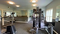 This fitness center offers a comprehensive workout environment designed to support a variety of exercise routines. The space is finished with a patterned low-pile carpet that defines the workout floor, while soft green-gray walls and white crown molding lend a clean, polished look to the interior. Flush-mount ceiling lights provide consistent, even illumination throughout the room, complementing the natural light that filters in through several windows along the walls. A large wall mirror expands the sense of space and allows users to monitor their form during exercise. The centerpiece of the room is a multi-station cable and strength training machine, suitable for a wide range of resistance exercises. Additional equipment is visible throughout, including a stability ball, a weight bench, and what appears to be cardio machines positioned along the perimeter. A half-wall partition with a wooden rail divides a secondary area, which may serve as a stretching or warm-up zone. Informational charts are posted on the walls, adding a functional, gym-oriented character to the space. Overall, the fitness center presents a well-organized and inviting atmosphere for residents or members looking to maintain an active lifestyle.