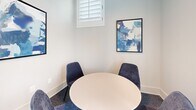 Located at 185 Marquis Way in Freeport, Florida, this single-floor coworking office is a thoughtfully designed workspace that balances functionality with contemporary style. The main area is anchored by a large rectangular conference table surrounded by plush navy blue chairs, positioned beneath a striking geometric pendant light fixture. Tall, shuttered windows flood the room with natural light while offering views of the surrounding greenery, creating a bright and inviting atmosphere. Textured blue-grey carpeting flows throughout the space, adding warmth and acoustic comfort underfoot. Along the perimeter, a row of individual workstations features sleek countertops, task chairs, and stylish gold elongated pendant lights, complemented by curated abstract artwork that lends a creative energy to the room. A semi-private nook with built-in booth seating, patterned upholstery, and a warm yellow accent ceiling offers a more intimate setting for focused work or casual conversation, complete with its own wall-mounted display. A small round table and seating area provides yet another collaboration option. The space is finished with clean white walls, black-framed doors, and modern lighting details throughout, resulting in a polished, professional environment well-suited for productivity and connection.