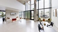 Located at 1402 Tacoma Ave S in Tacoma, Washington, this contemporary amenity space delivers a striking blend of industrial and modern design across a single, generously sized floor. The grand entrance lobby commands immediate attention with soaring double-height ceilings, floor-to-ceiling black-framed windows, and polished concrete floors that reflect abundant natural light flooding in from the street-facing glass facade. Exposed concrete columns and structural elements contrast elegantly with crisp white walls and a geometric, sculptural panel feature wall finished in soft sage green.

The lounge areas are thoughtfully appointed, with one featuring an exposed concrete accent wall, a built-in seating bench, and a curated mix of accent chairs arranged around patterned rugs. A separate, more intimate lounge showcases exposed concrete beam ceilings with acoustic paneling, generous sectional sofas, and direct views to an exterior glass-enclosed courtyard area. An open, light-filled activity zone benefits from a dramatic wall of windows, exposed coffered concrete ceilings, and polished floors, creating a bright and versatile gathering space. A dedicated media room, complete with acoustic ceiling tiles, multiple large-screen displays, and casual leather bean bag seating, provides an entertainment-focused retreat. Throughout, greenery, curated artwork, and warm textiles soften the industrial aesthetic.
