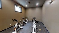 This single-floor fitness space is purpose-built for cycling workouts and group spin sessions. The room is outfitted with a lineup of True-brand stationary bikes arranged in a structured formation, providing ample space between each unit for comfortable use during individual or group exercise. The flooring features a durable, dark-toned carpet well-suited for a fitness environment, offering both cushioning and stability underfoot. The walls are finished in a warm taupe tone, creating a focused yet welcoming workout atmosphere. Recessed ceiling lighting ensures the room is well-lit throughout, while a ceiling fan positioned at the center promotes steady airflow during high-intensity sessions. A window along one wall allows for natural light to enter the space, adding a refreshing openness to the interior. The overall layout is clean and functional, with a no-frills design that keeps the emphasis firmly on fitness. This space is well-suited for facilities looking to offer a dedicated cycling or cardio area that is both practical and comfortable for regular use.