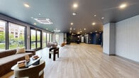 Located at 269 Orange Street in New Haven, CT, this thoughtfully designed amenity space offers a generous, single-floor layout that balances social gathering, dining, fitness, and relaxation. Light wood-look tile flooring flows continuously throughout, anchoring the space with a warm contrast against the dramatic dark ceilings and deep navy accent walls.

The open lounge area is furnished with contemporary seating arrangements and benefits from expansive floor-to-ceiling glass doors that open directly to an outdoor patio and green space, flooding the interior with natural light and offering seamless indoor-outdoor connectivity. Recessed lighting and ceiling-mounted HVAC units ensure year-round comfort.

A dedicated dining and kitchen zone features a long communal table, built-in wall ovens, ample cabinetry, and pendant lighting — ideal for resident gatherings or catered events. Adjacent to the lounge, a glass-walled fitness center provides a visually open yet defined workout area equipped with strength training machines.

Intimate seating alcoves with modern lounge chairs and side tables offer quieter retreats within the broader space. Textured concrete-finish walls add an architectural sophistication throughout. Additional private rooms, accessible via sleek grey doors, provide flexible utility spaces. The overall ambiance is contemporary, refined, and purposefully versatile.