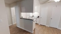 This single-floor space showcases a practical and well-organized layout centered around an open kitchen design. The kitchen is equipped with white cabinetry that extends to the upper walls, providing ample storage, while under-cabinet lighting illuminates the dark countertops below. A gas range and white appliances complement the clean, neutral color palette throughout. A peninsula with a dark countertop and integrated sink serves as a natural divider between the kitchen and the adjoining living area, offering additional workspace and a casual dining or prep area. The walls are painted in a light gray tone, contributing to a bright and airy feel, while light wood-style flooring runs continuously across the space, adding warmth and visual cohesion. A textured ceiling adds a classic touch to the interior. The layout is efficient and open, allowing for easy movement between functional zones. A white interior door near the kitchen provides access to an adjacent room or storage area, adding to the overall practicality of the space.