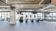 Located in downtown Salt Lake City, this well-appointed fitness center offers a generously sized, single-floor layout designed to accommodate a wide range of workout needs. The space is defined by its industrial-modern aesthetic, showcasing exposed concrete ceilings with visible ductwork and structural columns that lend an urban character throughout.

Floor-to-ceiling windows line multiple walls, flooding the facility with abundant natural light and providing sweeping views of the surrounding Salt Lake City skyline and mountains. The flooring transitions between durable rubber tiles and cork-style surfaces, each zone thoughtfully designed to suit its intended use.

The cardio section is well-equipped with treadmills, ellipticals, stair climbers, and stationary bikes, all positioned to take advantage of the panoramic city views. A dedicated strength training area features cable machines and Life Fitness multi-functional equipment, complemented by a free weights station. A functional training zone houses rowing machines, spin bikes, battle ropes, a tire for functional exercises, and open floor space ideal for stretching or dynamic workouts. Wall-mounted mirrors and multiple flat-screen televisions are distributed throughout the space. The facility also includes access to an outdoor area through glass doors, further enhancing the open, airy atmosphere of this comprehensive fitness destination.