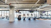 Located in downtown Salt Lake City, this well-appointed fitness center offers a generously sized, single-floor layout designed to accommodate a wide range of workout needs. The space is defined by its industrial-modern aesthetic, showcasing exposed concrete ceilings with visible ductwork and structural columns that lend an urban character throughout.

Floor-to-ceiling windows line multiple walls, flooding the facility with abundant natural light and providing sweeping views of the surrounding Salt Lake City skyline and mountains. The flooring transitions between durable rubber tiles and cork-style surfaces, each zone thoughtfully designed to suit its intended use.

The cardio section is well-equipped with treadmills, ellipticals, stair climbers, and stationary bikes, all positioned to take advantage of the panoramic city views. A dedicated strength training area features cable machines and Life Fitness multi-functional equipment, complemented by a free weights station. A functional training zone houses rowing machines, spin bikes, battle ropes, a tire for functional exercises, and open floor space ideal for stretching or dynamic workouts. Wall-mounted mirrors and multiple flat-screen televisions are distributed throughout the space. The facility also includes access to an outdoor area through glass doors, further enhancing the open, airy atmosphere of this comprehensive fitness destination.