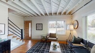 This two-floor home offers a thoughtfully designed layout highlighted by distinctive architectural character. The main living area immediately draws attention with its striking exposed wood beam ceiling, painted white to create an airy, open feel. Warm hardwood flooring runs throughout the main level, complementing the neutral color palette. Multiple windows allow natural light to fill the space, while a modern staircase with black metal railings leads to the upper level.

The living room provides generous space for seating arrangements and is anchored by a bold geometric area rug. The kitchen is a standout feature, showcasing two-tone cabinetry—white uppers paired with charcoal lowers—butcher block countertops, a farmhouse sink, subway tile backsplash, open shelving, and stainless steel appliances.

Both bedrooms are well-proportioned and feature ceiling fans, hardwood floors, and windows that allow ample natural light. One bedroom displays a darker, moody aesthetic with a black metal bed frame, while the second offers a lighter, serene ambiance. The shared bathroom includes a white vanity with a vessel-style sink, an oval mirror, and a tub-shower combination.