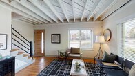 This two-floor home offers a thoughtfully designed layout highlighted by distinctive architectural character. The main living area immediately draws attention with its striking exposed wood beam ceiling, painted white to create an airy, open feel. Warm hardwood flooring runs throughout the main level, complementing the neutral color palette. Multiple windows allow natural light to fill the space, while a modern staircase with black metal railings leads to the upper level.

The living room provides generous space for seating arrangements and is anchored by a bold geometric area rug. The kitchen is a standout feature, showcasing two-tone cabinetry—white uppers paired with charcoal lowers—butcher block countertops, a farmhouse sink, subway tile backsplash, open shelving, and stainless steel appliances.

Both bedrooms are well-proportioned and feature ceiling fans, hardwood floors, and windows that allow ample natural light. One bedroom displays a darker, moody aesthetic with a black metal bed frame, while the second offers a lighter, serene ambiance. The shared bathroom includes a white vanity with a vessel-style sink, an oval mirror, and a tub-shower combination.