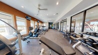 This fitness center offers a comprehensive workout environment thoughtfully arranged across a single, open floor. The space features a contemporary design with a striking combination of warm terracotta-orange accent walls and crisp white ceilings, creating an energizing atmosphere. Recessed lighting throughout, complemented by ceiling fans, ensures the space remains well-lit and comfortable during workouts.

Natural light fills the room through a series of windows along one wall, brightening the cardio zone, which is lined with a robust selection of treadmills, elliptical trainers, stair climbers, and stationary bikes—all Spirit-branded equipment mounted with personal display screens. Wall-mounted televisions above the windows provide additional entertainment for cardio users.

Toward the center and rear of the space, a functional training area houses a multi-station cable machine, a dumbbell rack, and adjustable weight benches, offering versatile strength training options. Kettlebells are neatly arranged along a rack on one side. Stability and exercise balls add a flexibility and core training dimension to the space.

A glass-walled room accessible through navy-framed double doors provides an enclosed area ideal for stretching, yoga, or private training sessions. The durable, rubberized flooring throughout supports high-impact activity while maintaining a clean, professional aesthetic. The layout is efficient and easy to navigate, making this fitness center a highly functional facility.