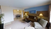 This single-floor residence showcases an open and functional layout that seamlessly connects the living and dining areas. The living room features a rich brown sofa set arranged around a sleek, dark rectangular coffee table with metal legs, all resting on plush wall-to-wall carpeting. A distinctive two-tone accent wall — combining a cool blue upper section with a warm tan lower section — serves as a visual focal point, complemented by a framed cityscape print that adds a cosmopolitan touch. A table lamp provides warm ambient lighting to the seating area.

The adjoining dining area is finished with smooth flooring and features a dark dining table surrounded by matching chairs, accented by decorative wall art. A ceiling light fixture illuminates the space, and the nearby kitchen is visible in the background, featuring white cabinetry and countertops. The neutral white ceiling and walls throughout the space create a bright, clean aesthetic that ties the open-plan layout together cohesively, making the residence feel welcoming and well-organized.