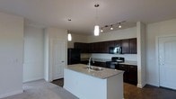 This single-floor space showcases a well-appointed kitchen as its centerpiece. The kitchen is defined by rich, dark espresso cabinetry that extends to the upper walls, providing generous storage throughout. A central island with a white base and speckled granite countertop serves as a functional focal point, equipped with a double-basin sink and a modern faucet. Matching granite countertops line the perimeter of the kitchen, complementing the dark cabinetry seamlessly. Black appliances, including a refrigerator, range, and built-in microwave, contribute to a cohesive, contemporary aesthetic. Overhead lighting is thoughtfully layered, combining pendant lights suspended above the island with a track lighting system that illuminates the cooking area effectively. The flooring transitions from what appears to be hardwood or tile in the kitchen to carpeting in the adjacent living area, visually distinguishing the two zones. Neutral, light-toned walls keep the space feeling open and bright. A paneled door off the kitchen provides access to an additional room or utility area, adding to the overall functionality of the layout.