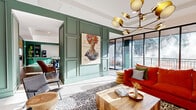 Located at 730 1/2 N First St in Minneapolis's vibrant North Loop neighborhood, this generously sized single-floor amenity and lobby space offers a thoughtfully designed environment that blends contemporary style with welcoming functionality. The space is unified by a striking sage and forest green color palette, expressed through detailed panel wainscoting and millwork that frames each zone with architectural elegance.

The main lounge area anchors the space with a bold red sectional sofa, eclectic woven accent chairs, and a live-edge wood coffee table, all set atop a graphic patterned area rug. A sculptural gold multi-arm chandelier draws the eye upward, while floor-to-ceiling black-framed windows flood the room with natural light and offer views of the surrounding streetscape.

The communal kitchen and dining area features deep green cabinetry with open shelving, a diamond-tile backsplash, a rolling library ladder, and a substantial dining table with designer chairs — ideal for gathering and collaboration.

Wide, light-filled hallways lined with curated gallery walls and warm wood ceiling accents connect the zones seamlessly. A secondary corridor leads to elevator bays set against stone-clad walls, providing a clean and functional transition point. Throughout the space, original artwork and lush greenery add warmth and personality to every corner.