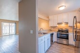This single-floor residence offers a well-organized layout that connects the kitchen, living area, and private rooms through a practical, open design. The kitchen is equipped with stainless steel appliances, including a refrigerator, electric range with a under-cabinet range hood, and dishwasher. White shaker-style cabinetry provides ample storage, complemented by speckled laminate countertops and a single-basin sink. A raised bar counter separates the kitchen from the adjoining living area, which receives natural light through a sliding glass door visible in the background.

The living area is generously proportioned, featuring light wood-style flooring that extends throughout the unit, warm beige walls, and track lighting on the ceiling. A hallway leads to the bedroom and bathroom. The bedroom is a quiet, well-lit space with a window fitted with vertical blinds, allowing natural light while maintaining privacy. The bathroom includes a full tub-shower combination, a toilet, a single vanity with a speckled countertop, and a small privacy window positioned high on the wall. Textured walls and consistent warm-toned paint unify the overall aesthetic of the space.